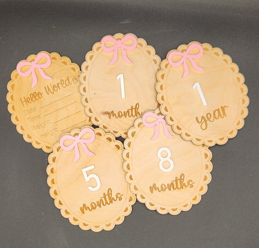 A set of wooden baby milestone cards with a light pink bow and white number on each, indicating various ages from 1 month to 1 year.