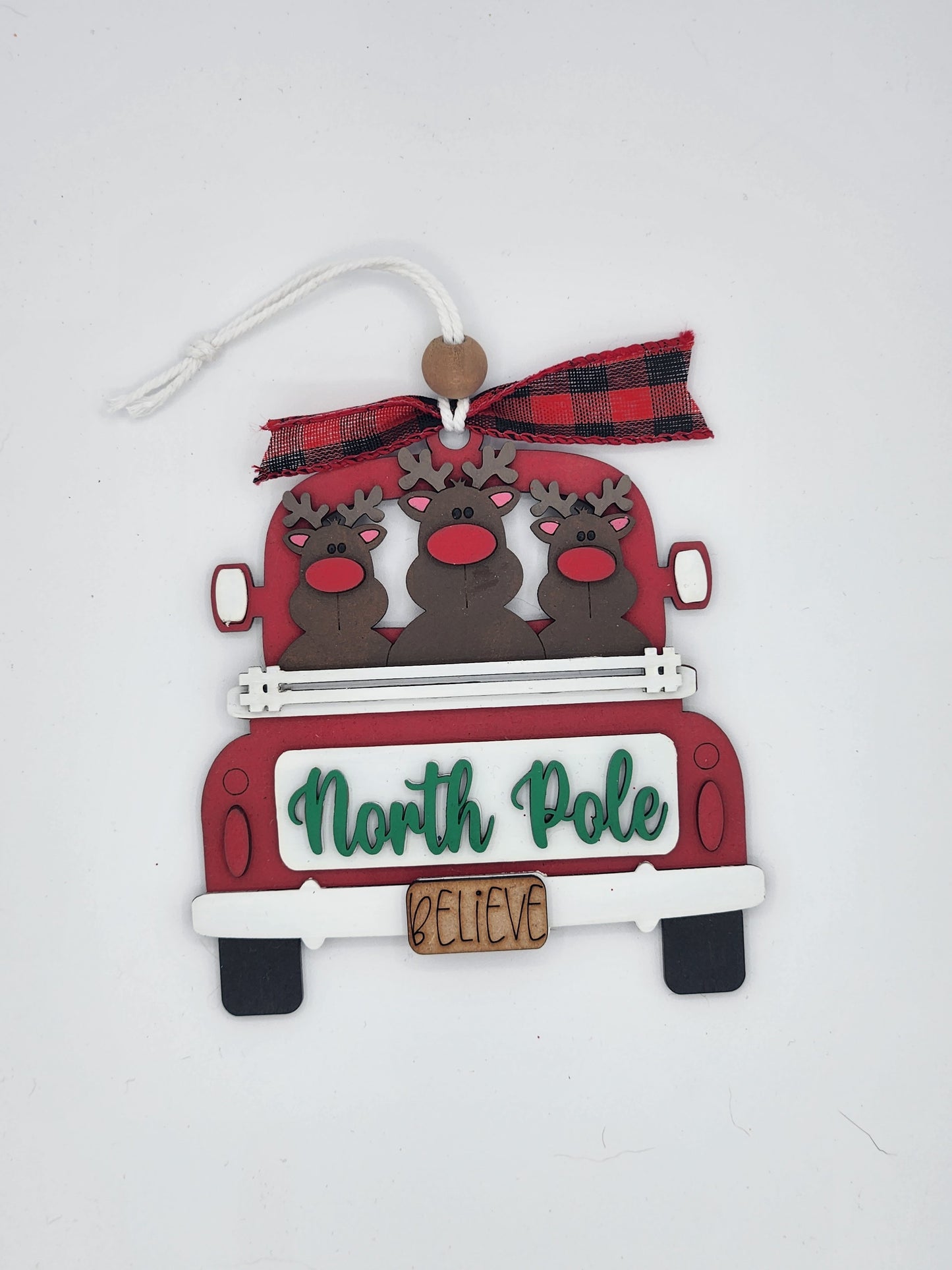 Decorative Christmas ornament with reindeer in a red truck and 'North Pole Believe' text on a white background.
