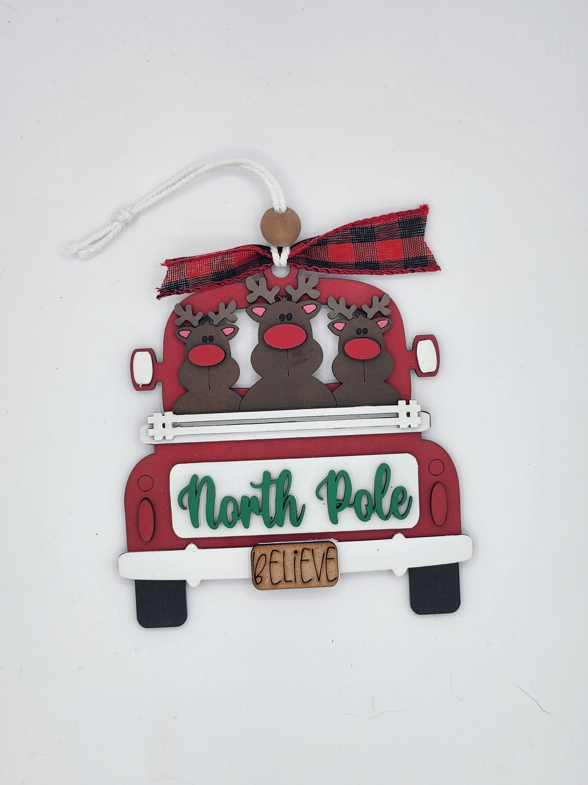 Decorative Christmas ornament with reindeer in a red truck and 'North Pole Believe' text on a white background.