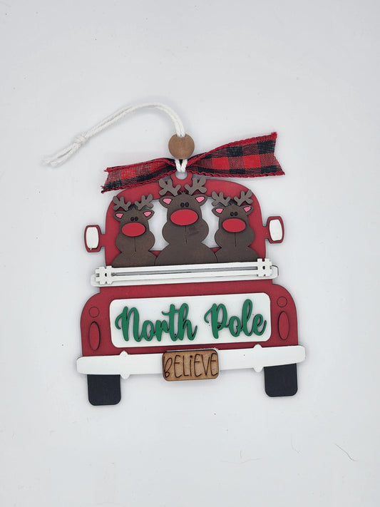 Decorative Christmas ornament with reindeer in a red truck and 'North Pole Believe' text on a white background.
