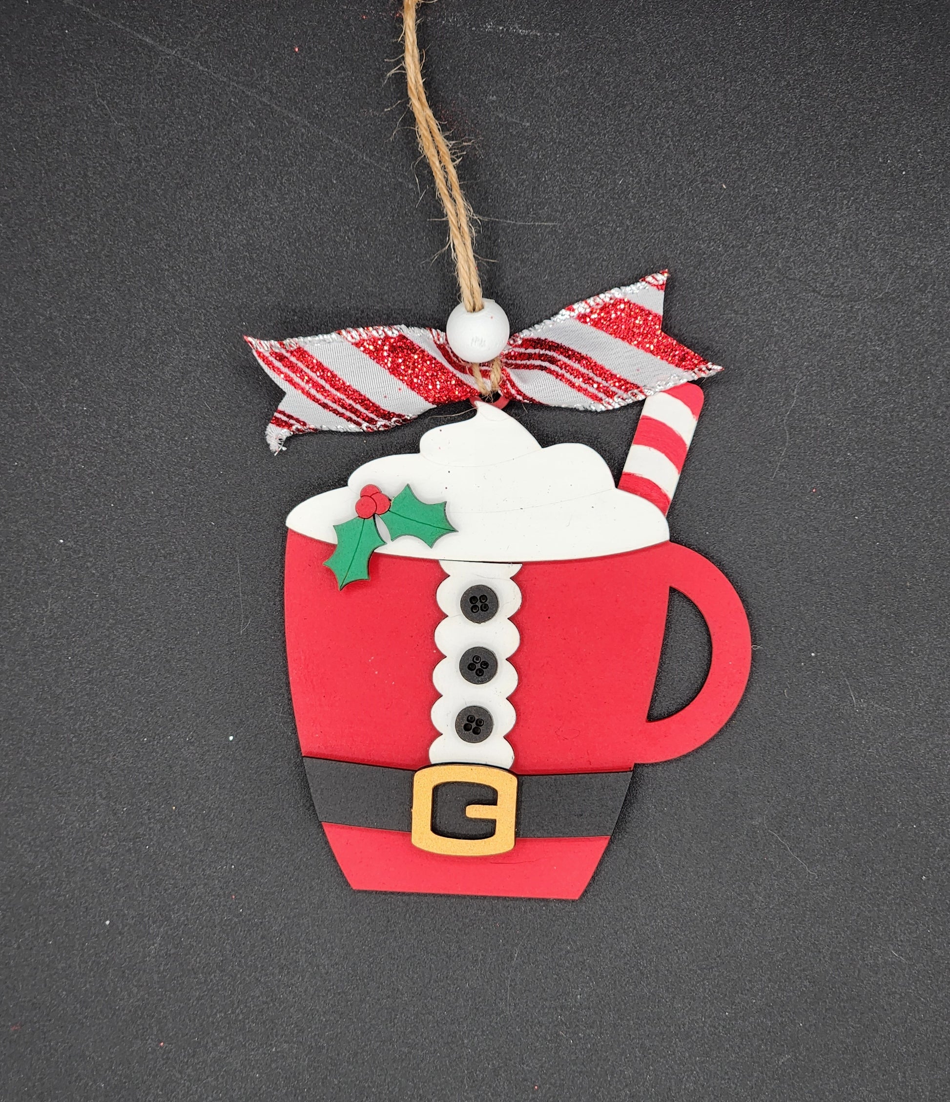 A DIY ornament depicting a Santa Claus outfit hot cocoa mug with a red cup of hot cocoa, white cream, and candy cane stripes on the ribbon.
