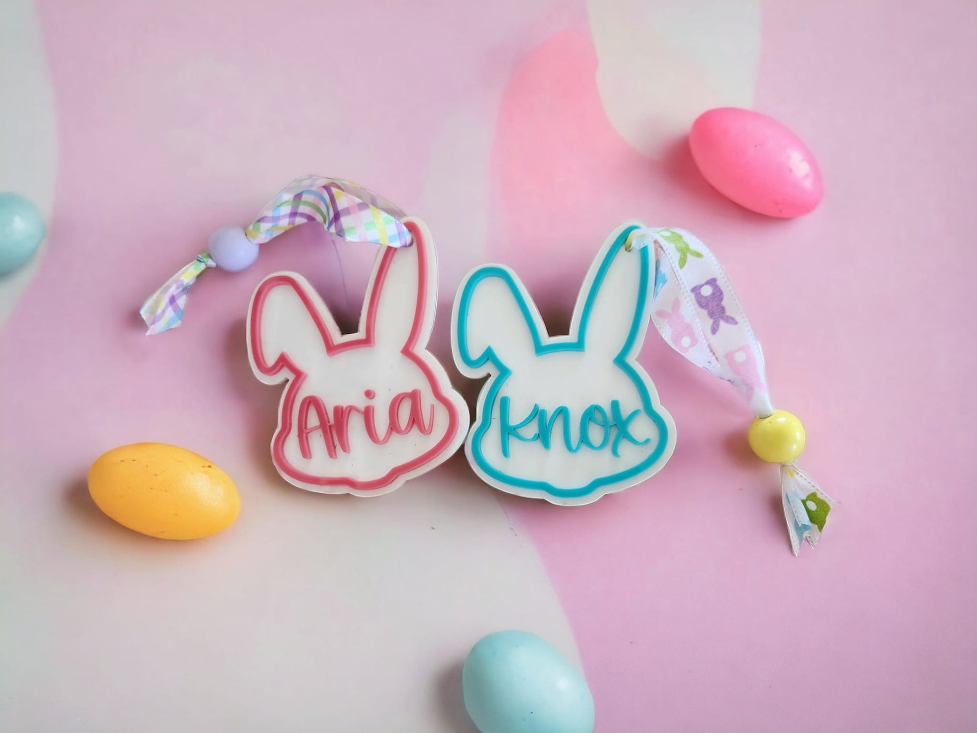 Customizable Easter name tags in the shapes of rabbits, with the names 'Aria' and 'Knox' written on them, surrounded by colorful candies and a pink background.