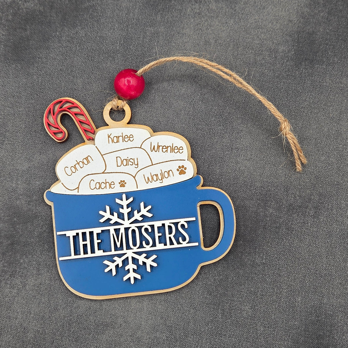 A personalized holiday ornament shaped like a blue mug filled with hot cocoa, featuring the name 'THE MOSERS' in white letters, surrounded by custom names in the cocoa foam, with a red bead and jute hanging string on a dark gray textured background.