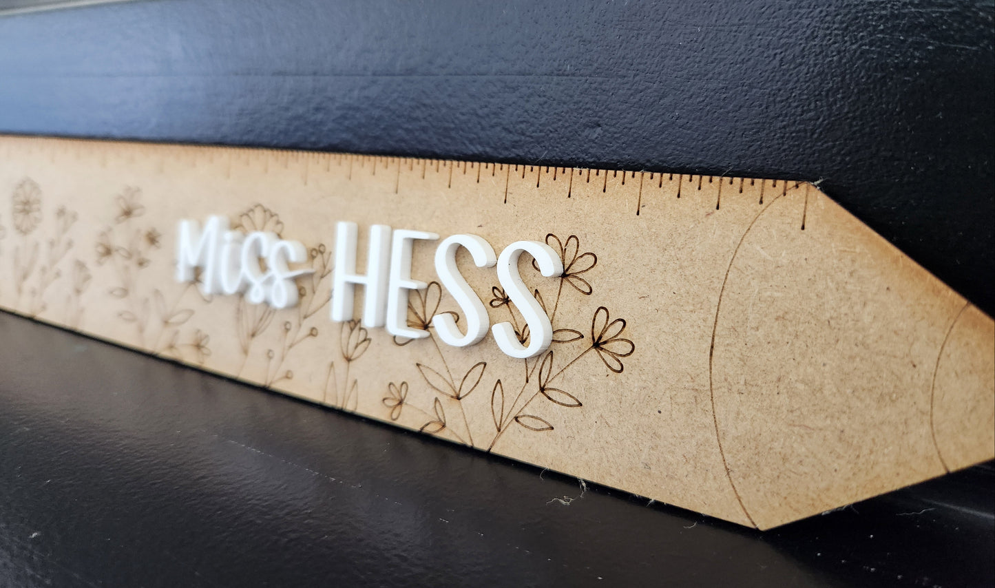 A personalized decorative ruler with the name 'Miss Hess' printed on it, alongside floral designs and measurement markings.