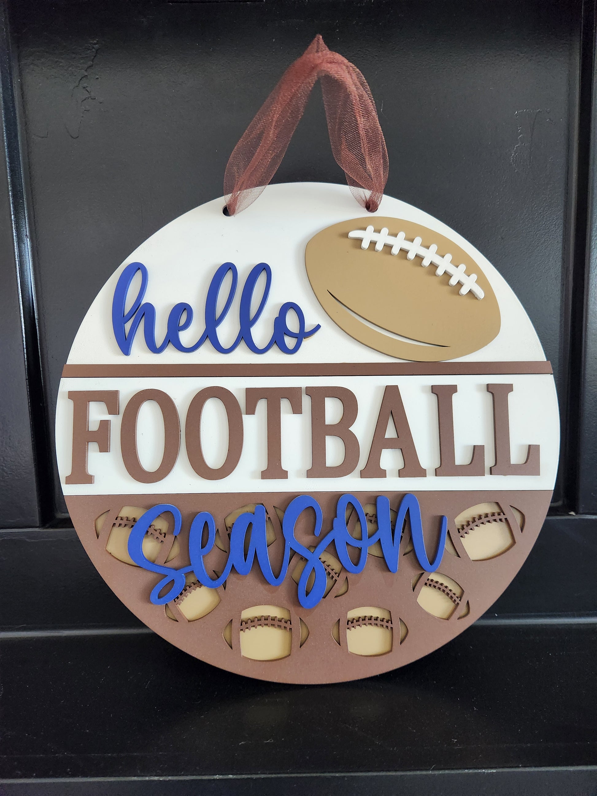 A round wooden door hanger with the phrase 'hello football season' painted in blue, brown, and beige colors, featuring footballs and a ribbon for hanging.
