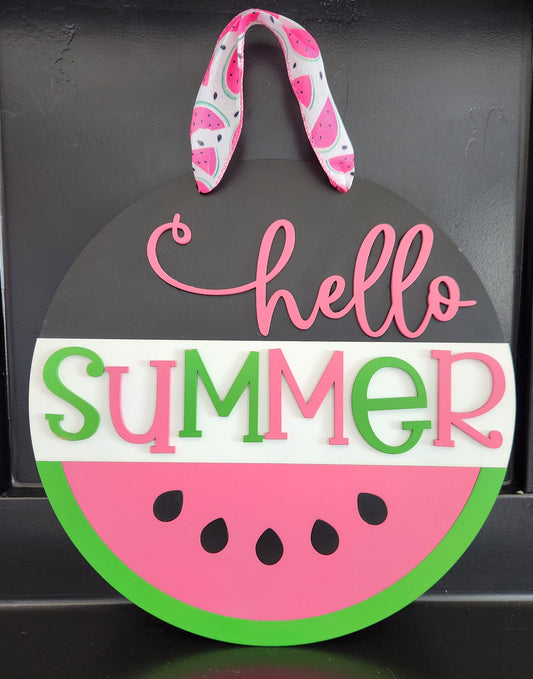 A round decorative sign with a watermelon design and the text 'hello SUMMER' in bold letters. The sign is hung by a ribbon at the top.