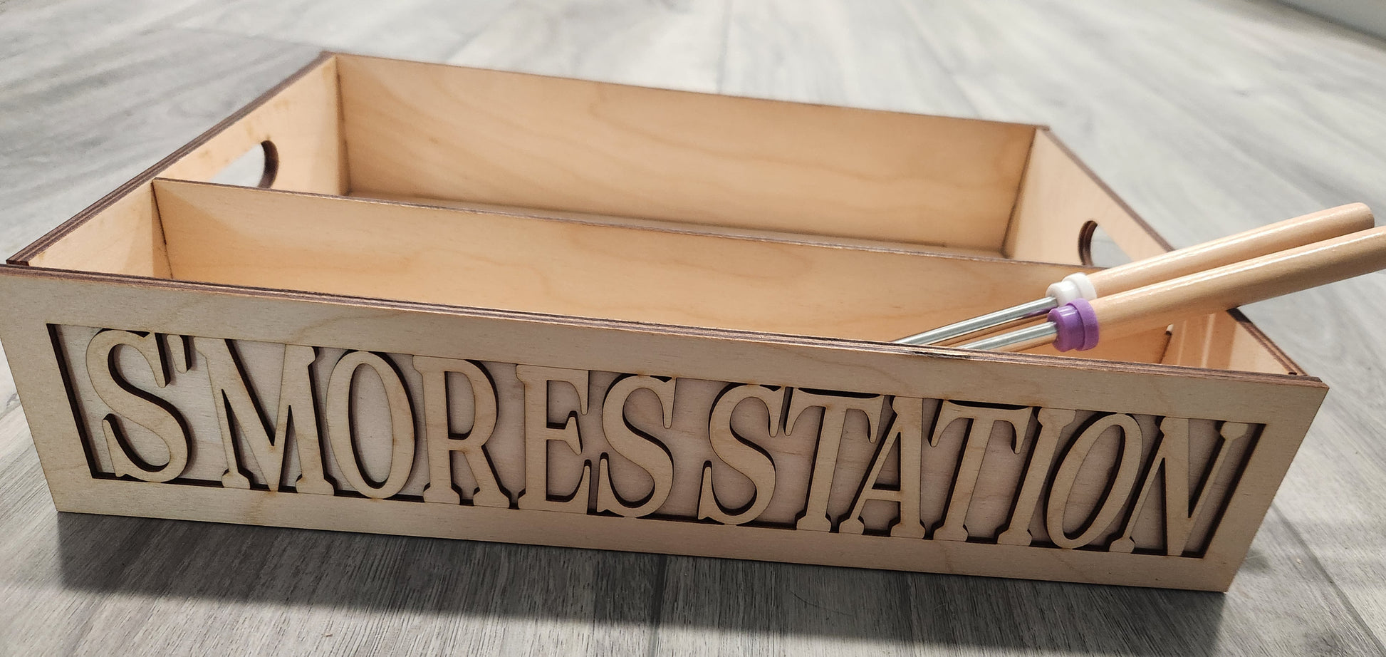 A wooden s'mores station box with 'SMORES STATION' cut out on the side, accompanied by two smores sticks with purple and white handles.