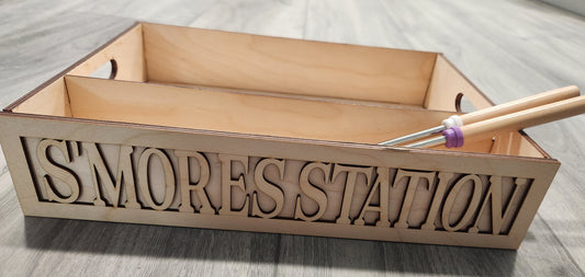 A wooden s'mores station box with 'SMORES STATION' cut out on the side, accompanied by two smores sticks with purple and white handles.