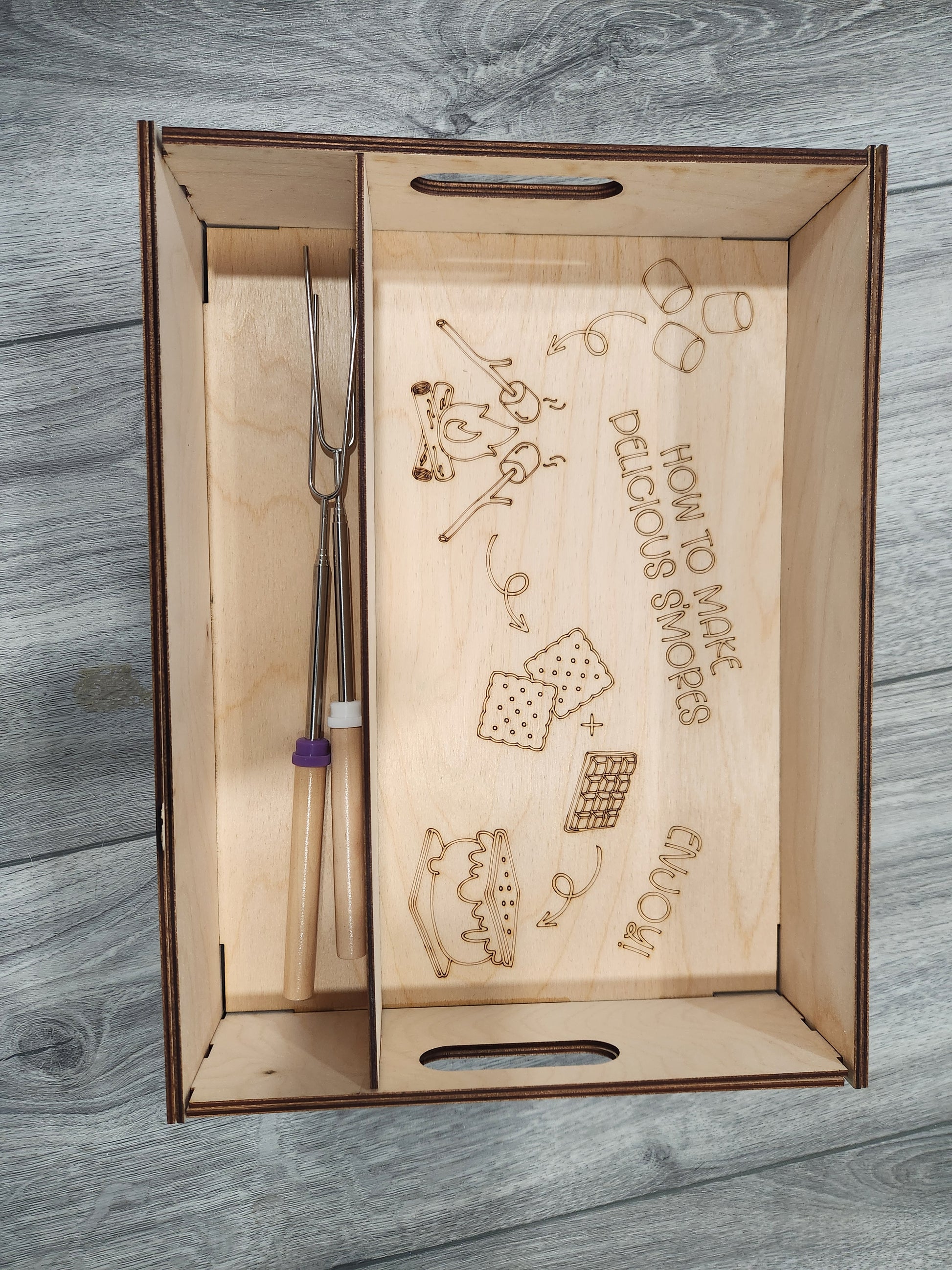 Wooden tray with engraved smores instructions and marshmallow roasting sticks on a wooden surface.