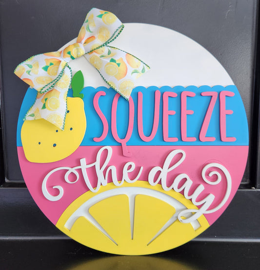 A 16-inch round decorative plaque with a lemon and a bow design, featuring the phrase 'Squeeze the day' in blue and pink colors.