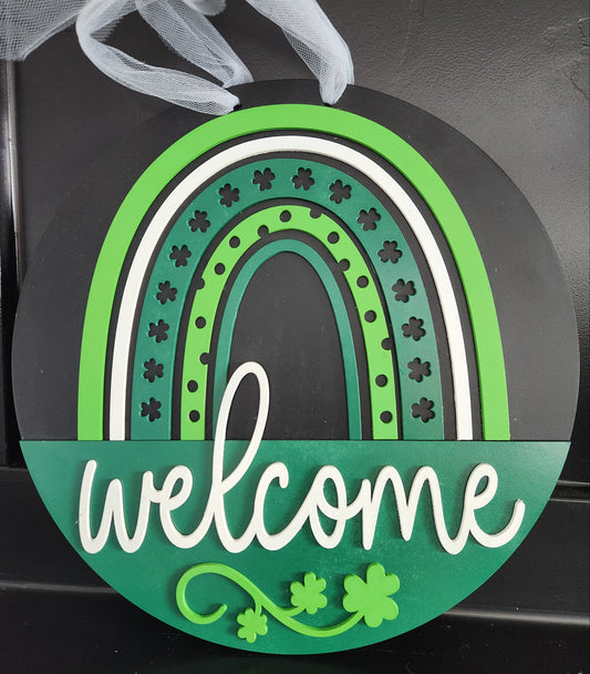 A round decorative door hanger with a green and black color scheme featuring a clover pattern and the word 'welcome' in the center, adorned with a white ribbon at the top.