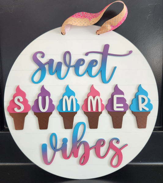 A circular wooden sign with the phrase 'Sweet Summer Vibes' painted in multi-colored letters, featuring ice cream cone shapes, with a ribbon at the top.