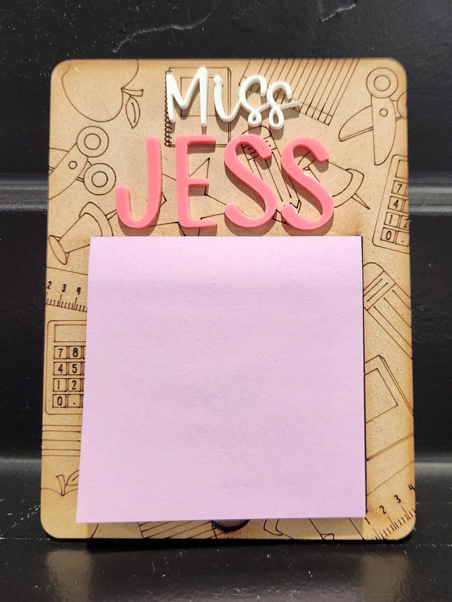 A personalized sticky note holder with a wooden base and a purple sticker on the front. The name 'Jess' is printed on the holder.