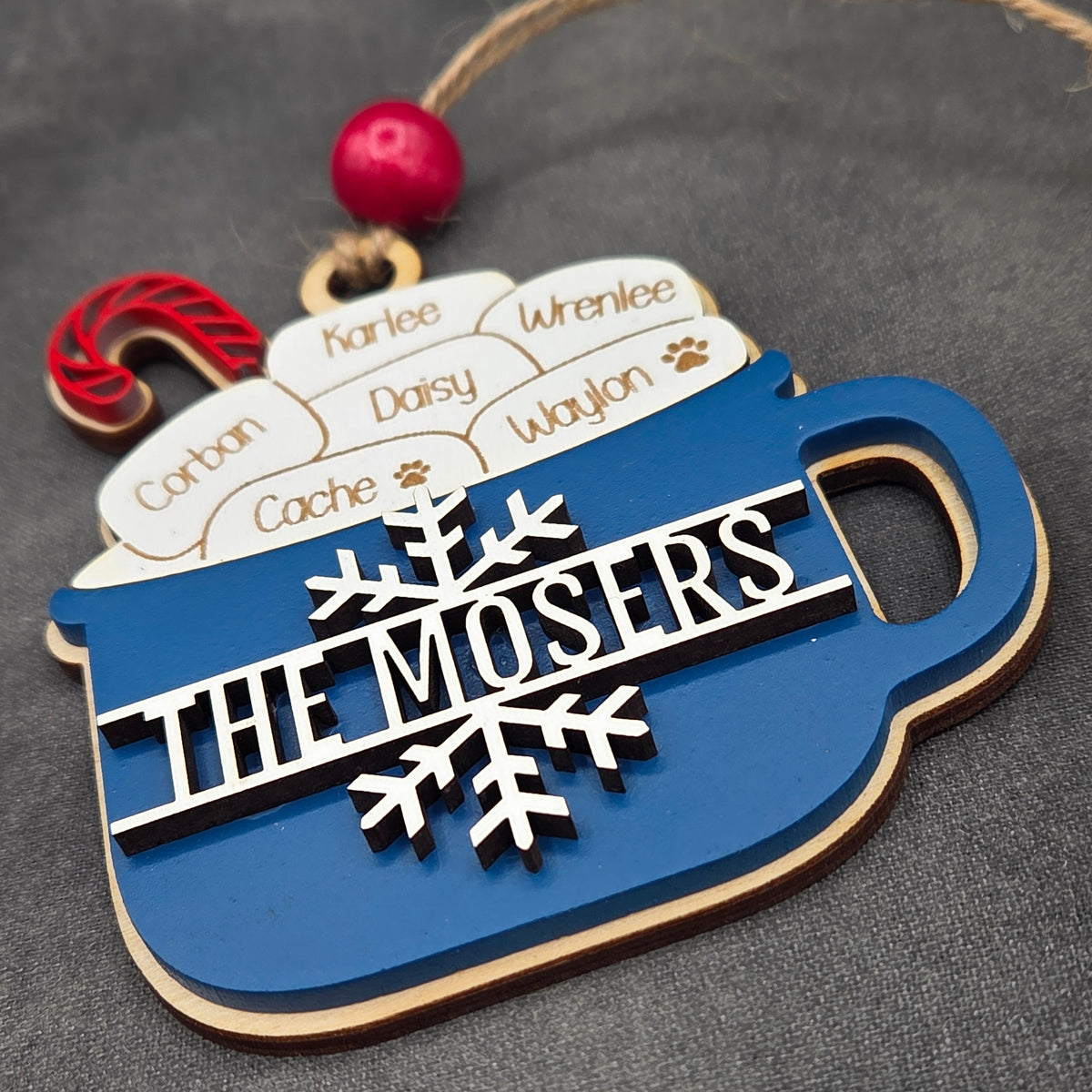 A wooden holiday ornament shaped like a blue mug filled with white marshmallows labeled with names, featuring a snowflake and the text 'THE MOSERS' in white. A red bead and string are attached at the top, with a small red candy cane accent on the left, photographed on a dark gray textured surface.